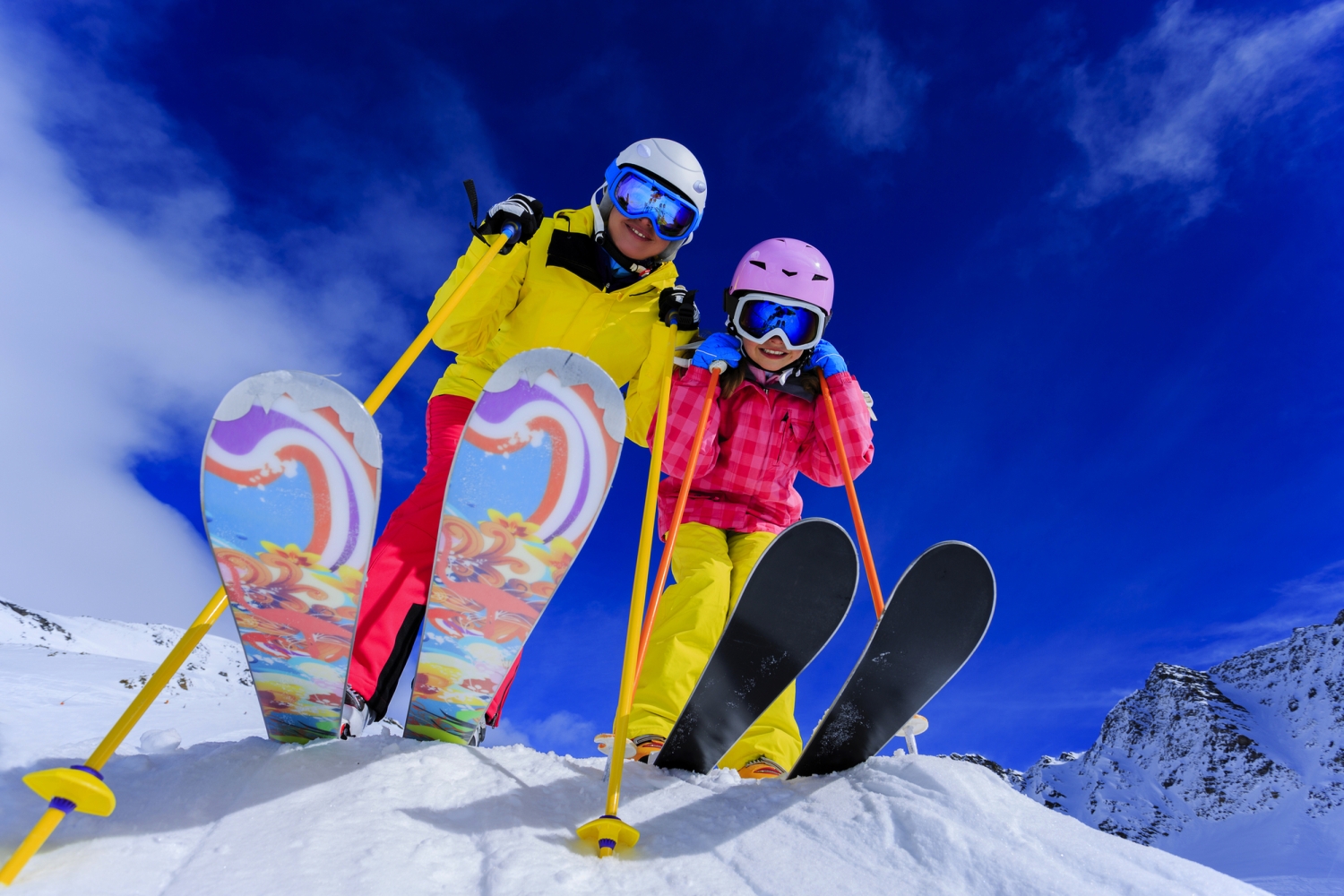 Zwei Kinder in bunter Skikleidung stehen auf einem verschneiten Hang mit Skiern.