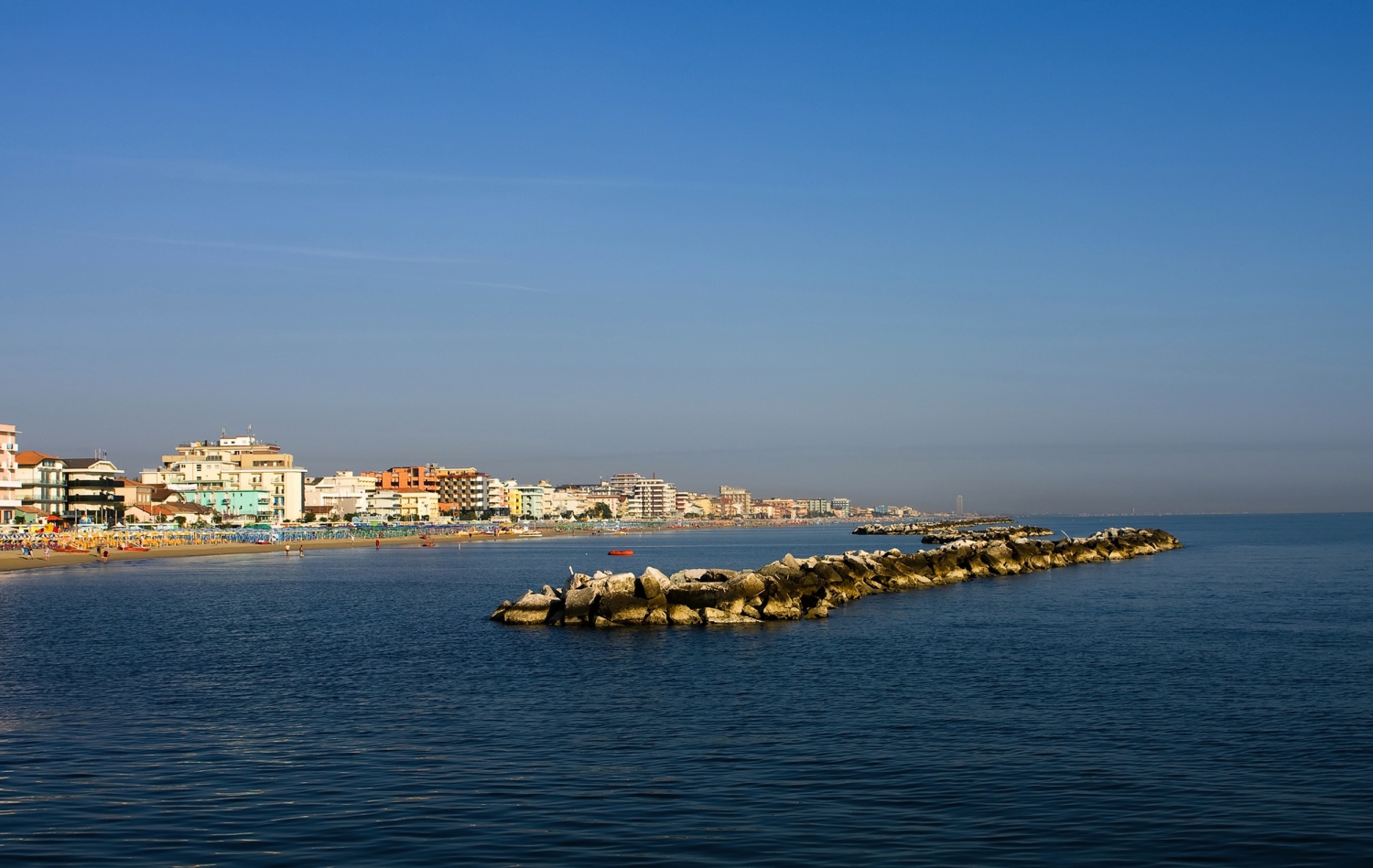 Strand und Gebäude entlang eines ruhigen Meeres mit Felsbarriere im Vordergrund.