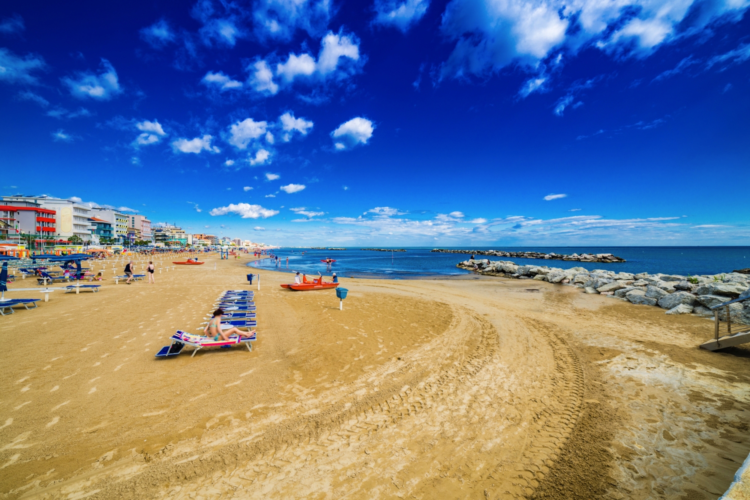 Sandstrand mit Liegestühlen, blauem Himmel und Meer, gesäumt von Gebäuden.
