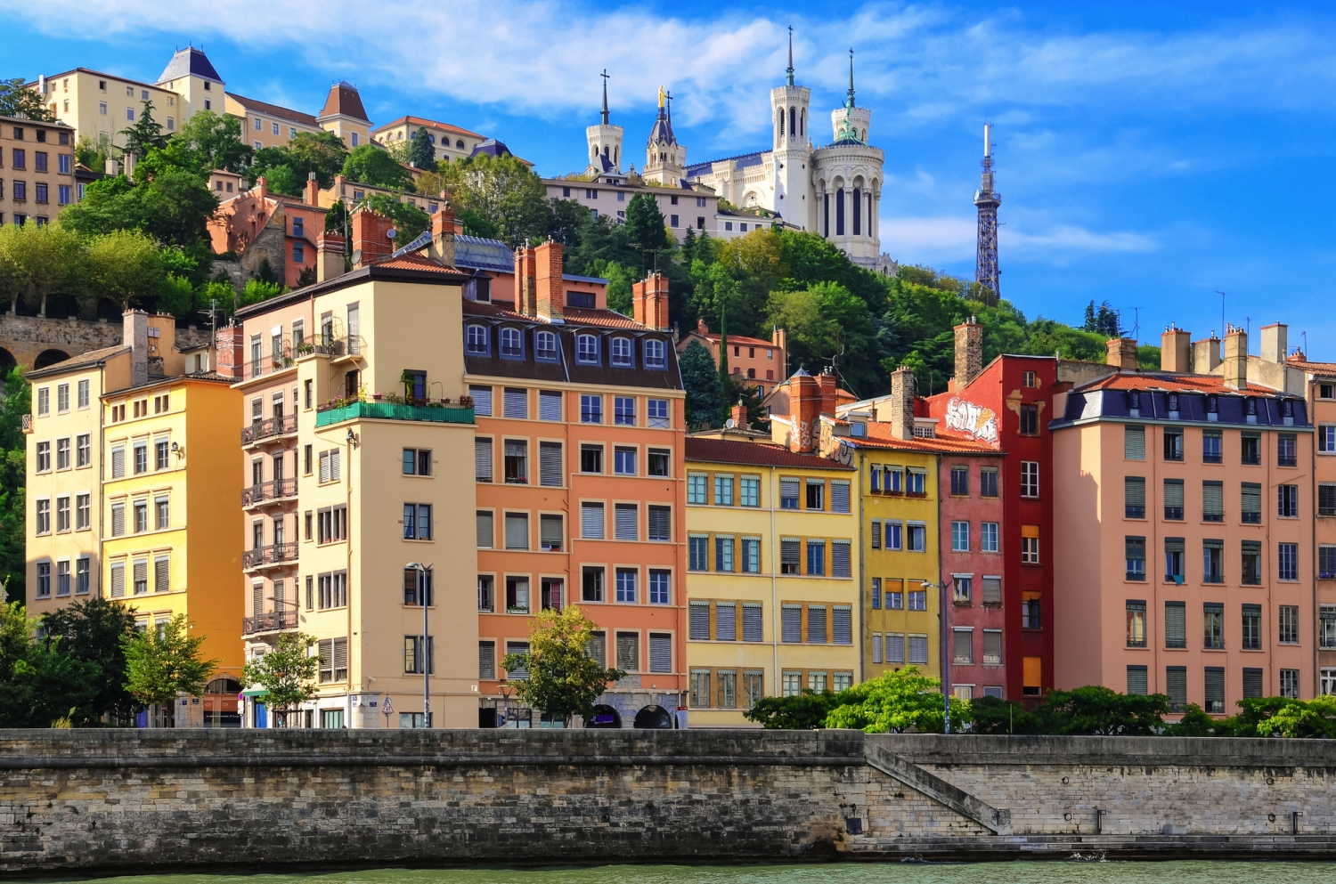 Bunte Gebäude am Flussufer mit Hügel und Kirche im Hintergrund.
