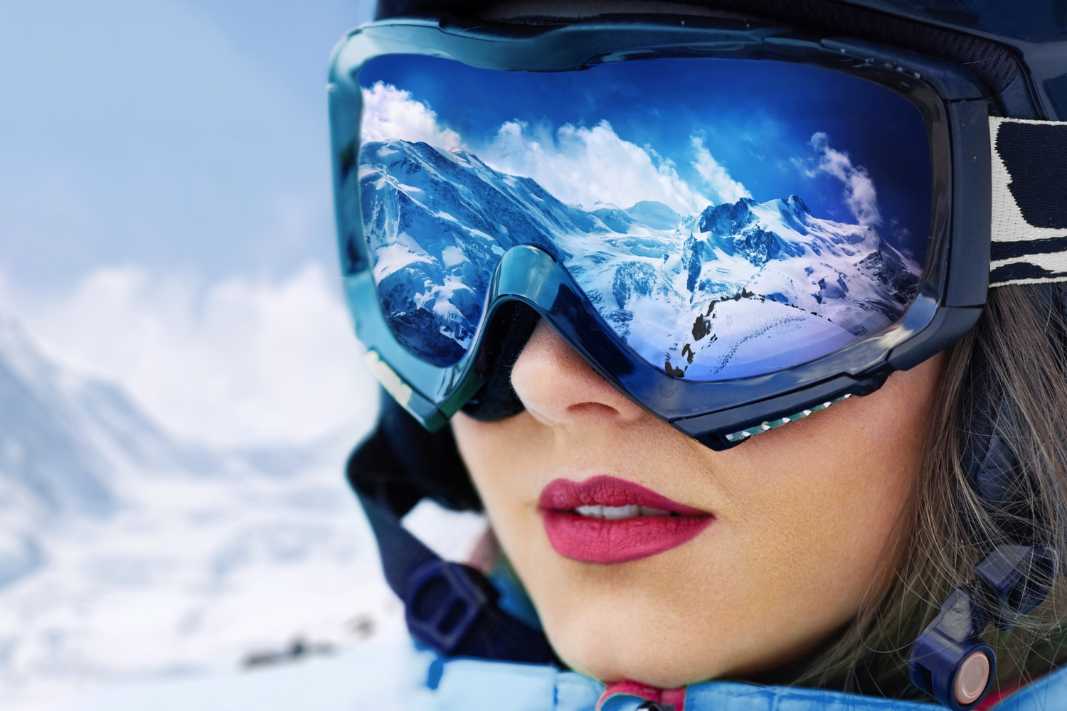 Eine Frau in Skibekleidung mit einer Skimaske, die Berge und blauen Himmel reflektiert.