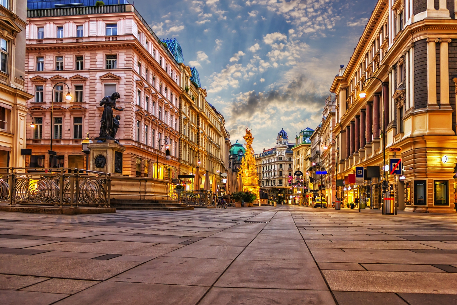 Prachtstraße mit historischen Gebäuden und einer Barocksäule bei Sonnenuntergang in der Stadt.