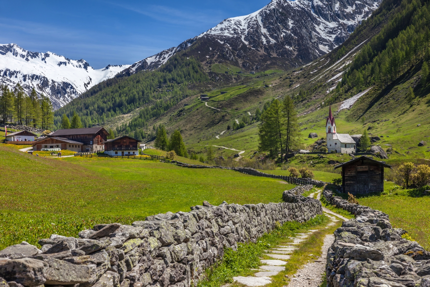 Berglandschaft mit Kirche, Wiesen, Steinweg und schneebedeckten Bergen im Hintergrund.