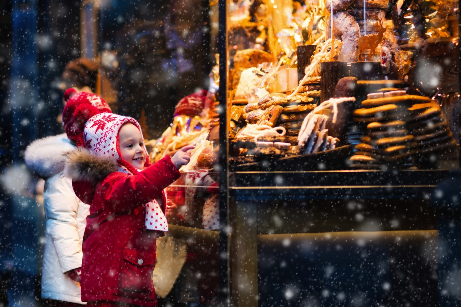 © famveldman - stock.adobe.com Kind im roten Mantel zeigt auf Lebkuchenauslage hinter einem Schaufenster im Schneefall.