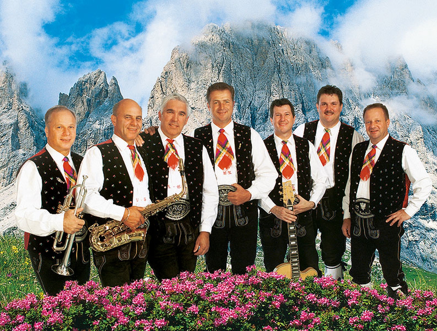 Sieben Männer in traditioneller Tracht vor Bergkulisse mit Alpenblumen.