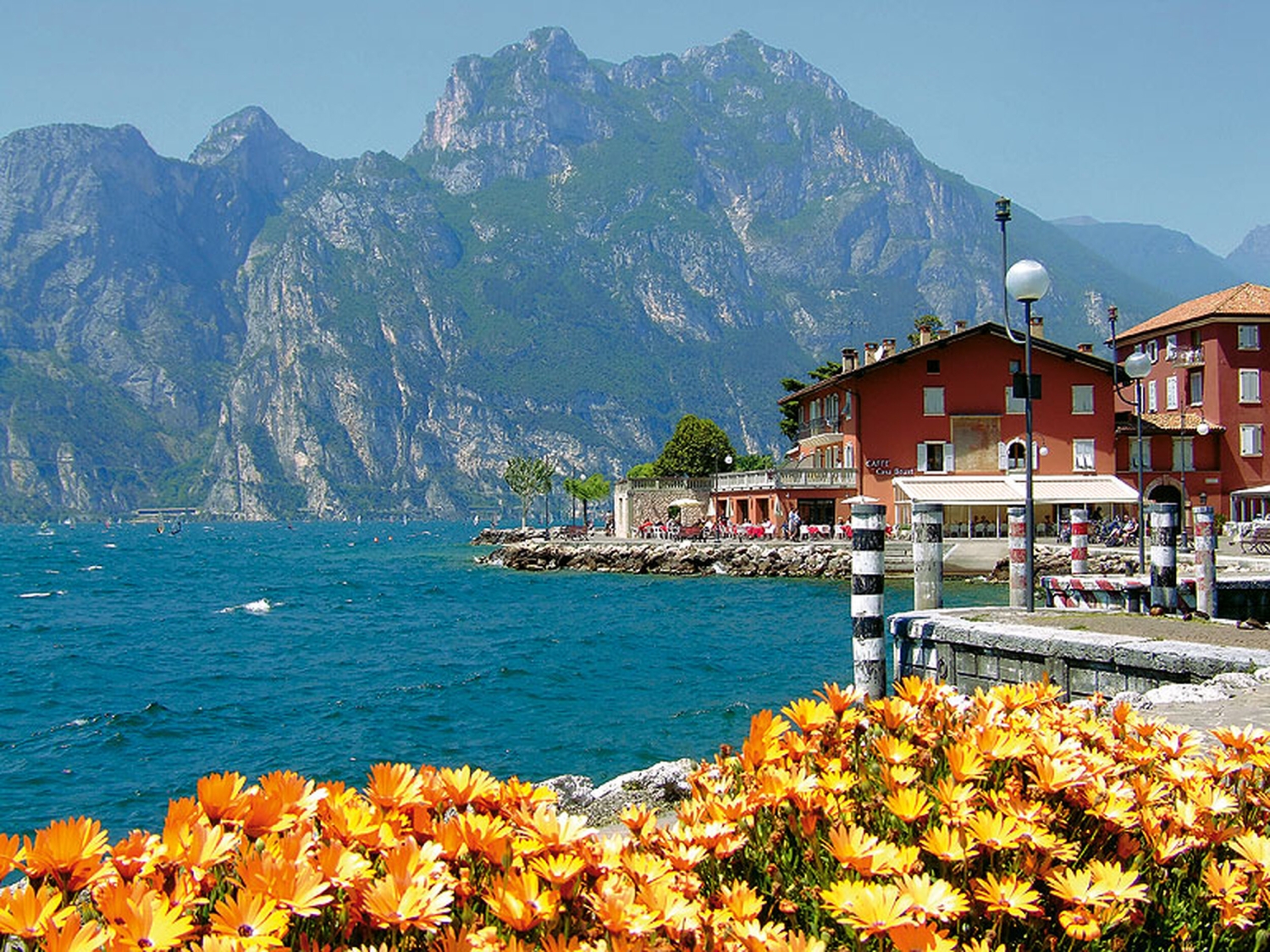 Gebäude am Ufer des Gardasees in Torbole mit Blumen im Vordergrund und Bergen im Hintergrund.