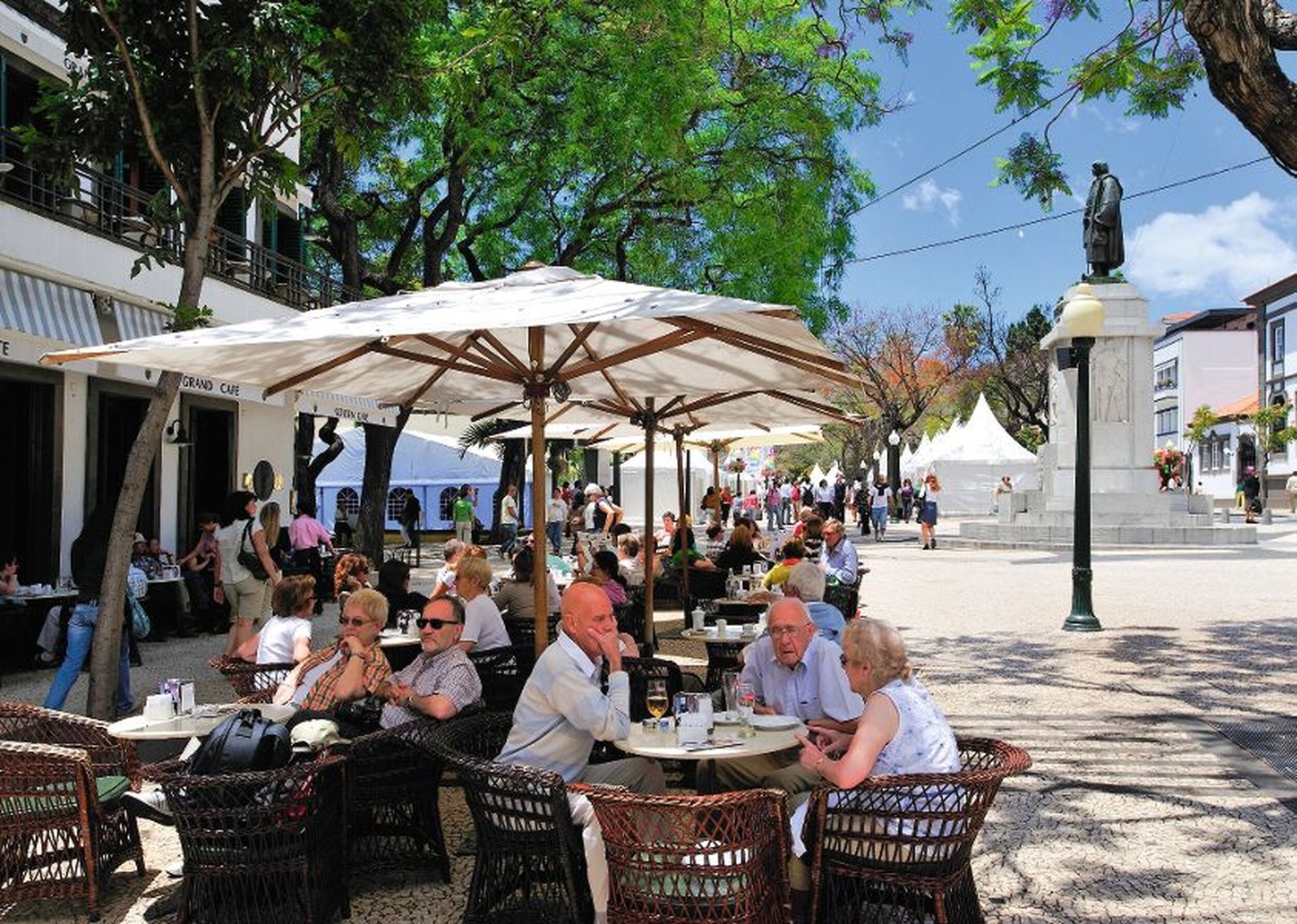 Ein belebtes Straßencafé in der Altstadt mit vielen Menschen, die unter Sonnenschirmen an Tischen sitzen und sich unterhalten, umgeben von grünen Bäumen und historischen Gebäuden.