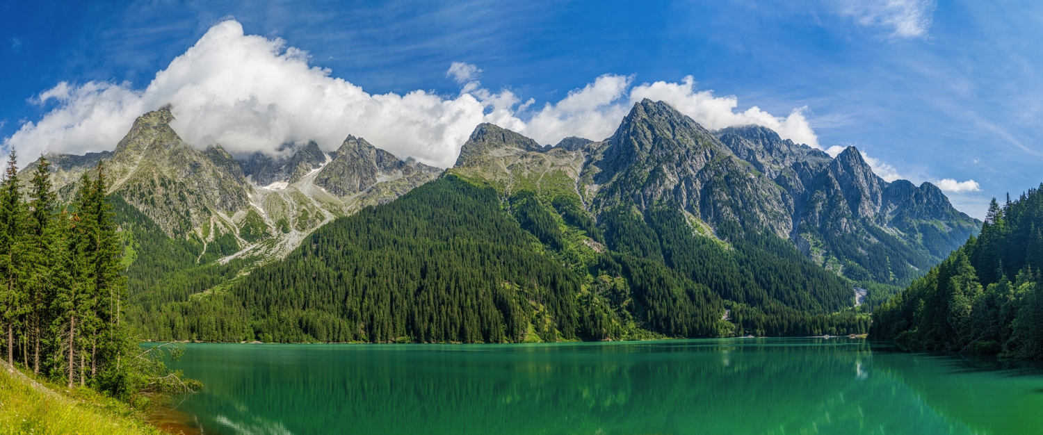 Bergsee mit grüner Wasserfläche und bewaldeten Bergen im Hintergrund bei klarem Himmel.