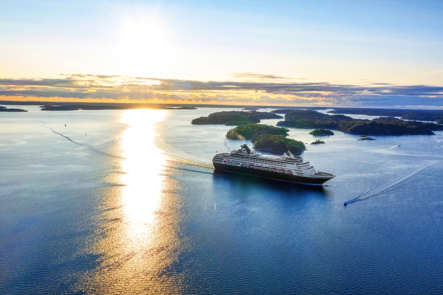 © nicko cruises Schiffsreisen GmbH Kreuzfahrtschiff auf ruhiger See bei Sonnenuntergang, umgeben von kleinen Inseln.