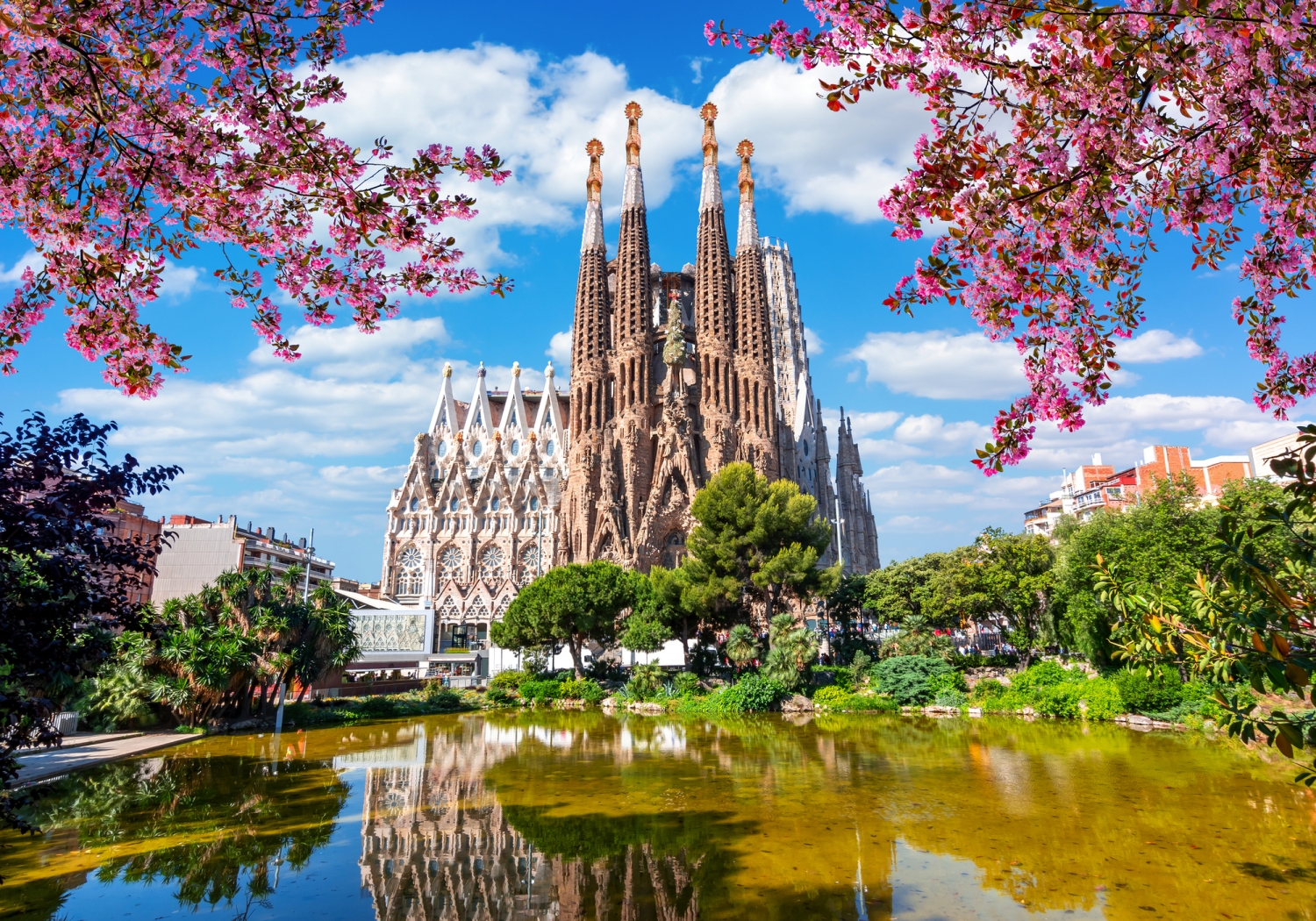 Sagrada Família in Barcelona, umgeben von blühenden Bäumen und Wasserreflexionen.