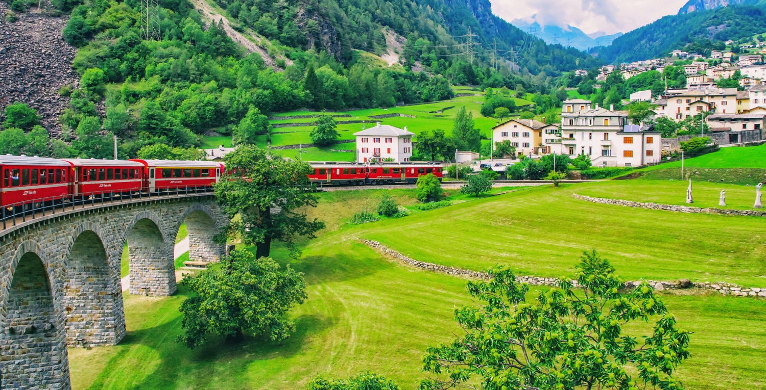 Roter Zug fährt über Steinbrücke in grüner alpiner Landschaft.