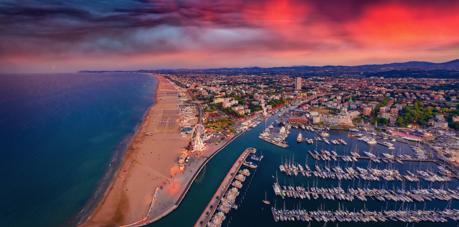 Küstenstadt mit Hafen und Sandstrand bei Sonnenuntergang aus der Luftperspektive.