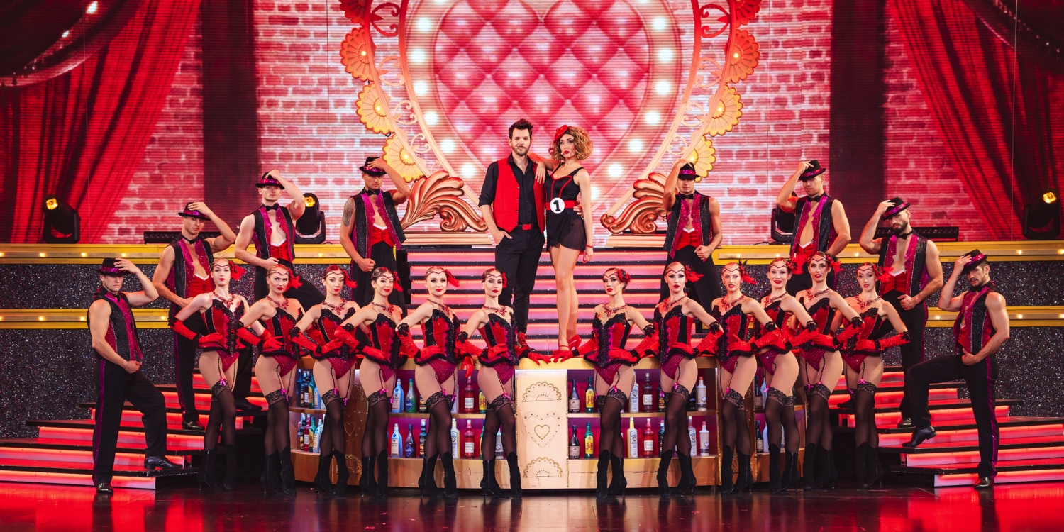 Eine Vielzahl von Tänzern in roten Kostümen auf einer Bühne mit Herzdekoration.