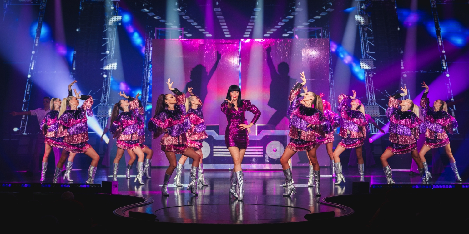 Eine Gruppe von Tänzerinnen in lila Kostümen auf einer Bühne mit bunten Lichtern.