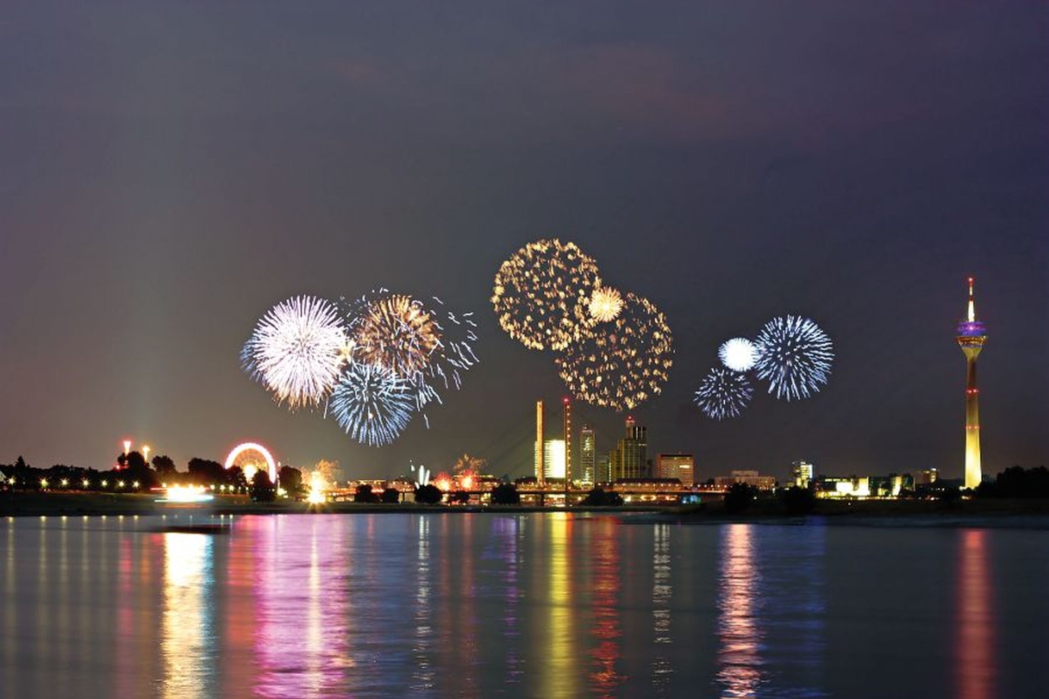 Feuerwerk über der Skyline von Düsseldorf mit dem Rheinturm und Reflexionen im Wasser.