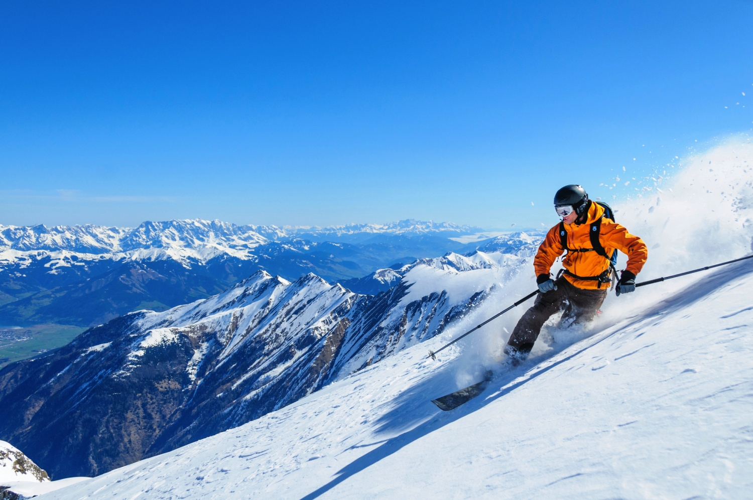 Skifahrer in orange Jacke fährt bergab auf verschneitem Hang mit Bergpanorama im Hintergrund.
