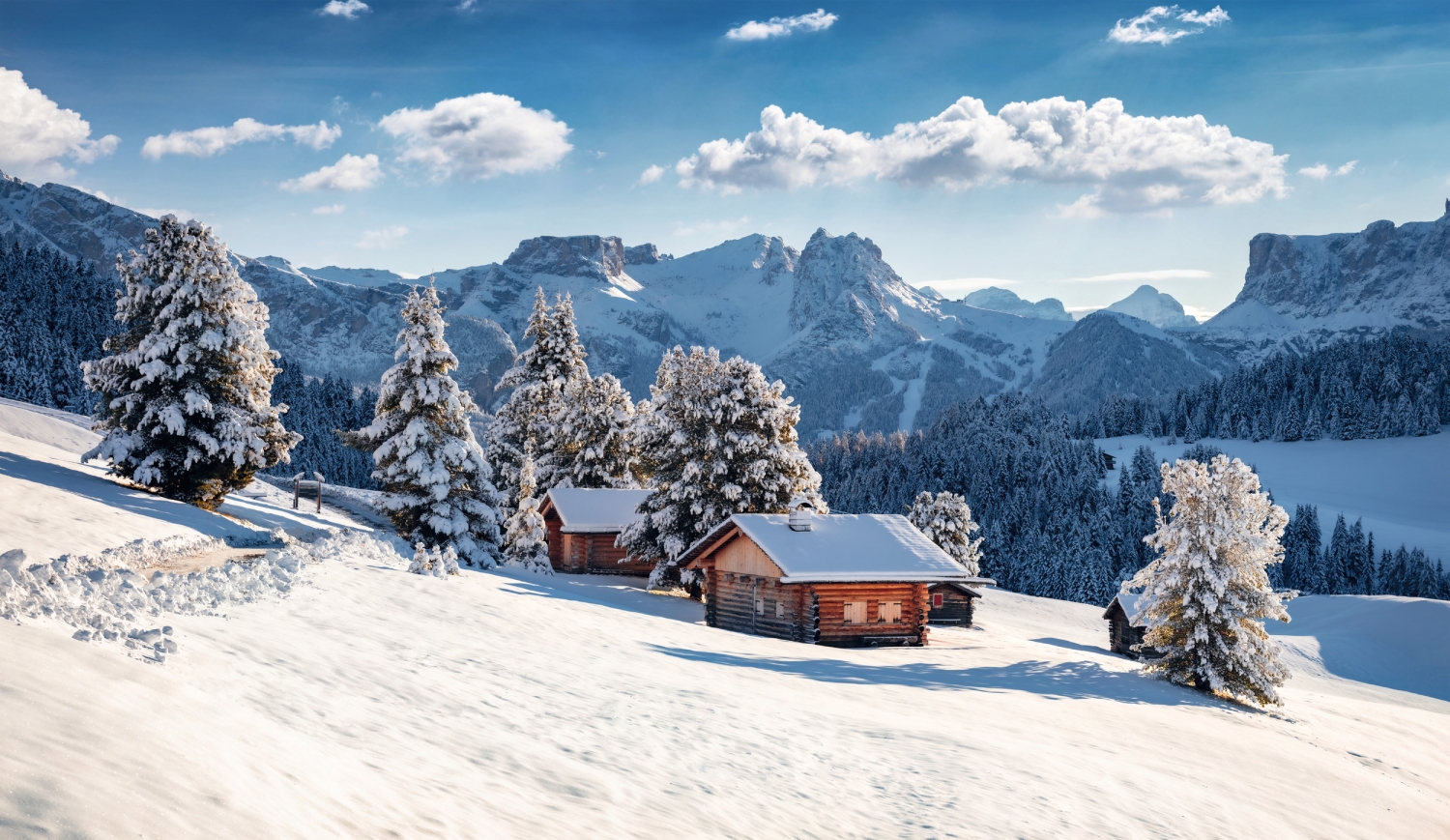 Verschneite Hütten in einer winterlichen Berglandschaft mit Tannenbäumen und strahlend blauem Himmel.