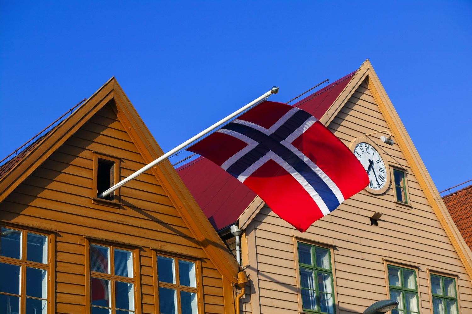 Norwegische Flagge an Holzhäusern mit Uhr und blauem Himmel.