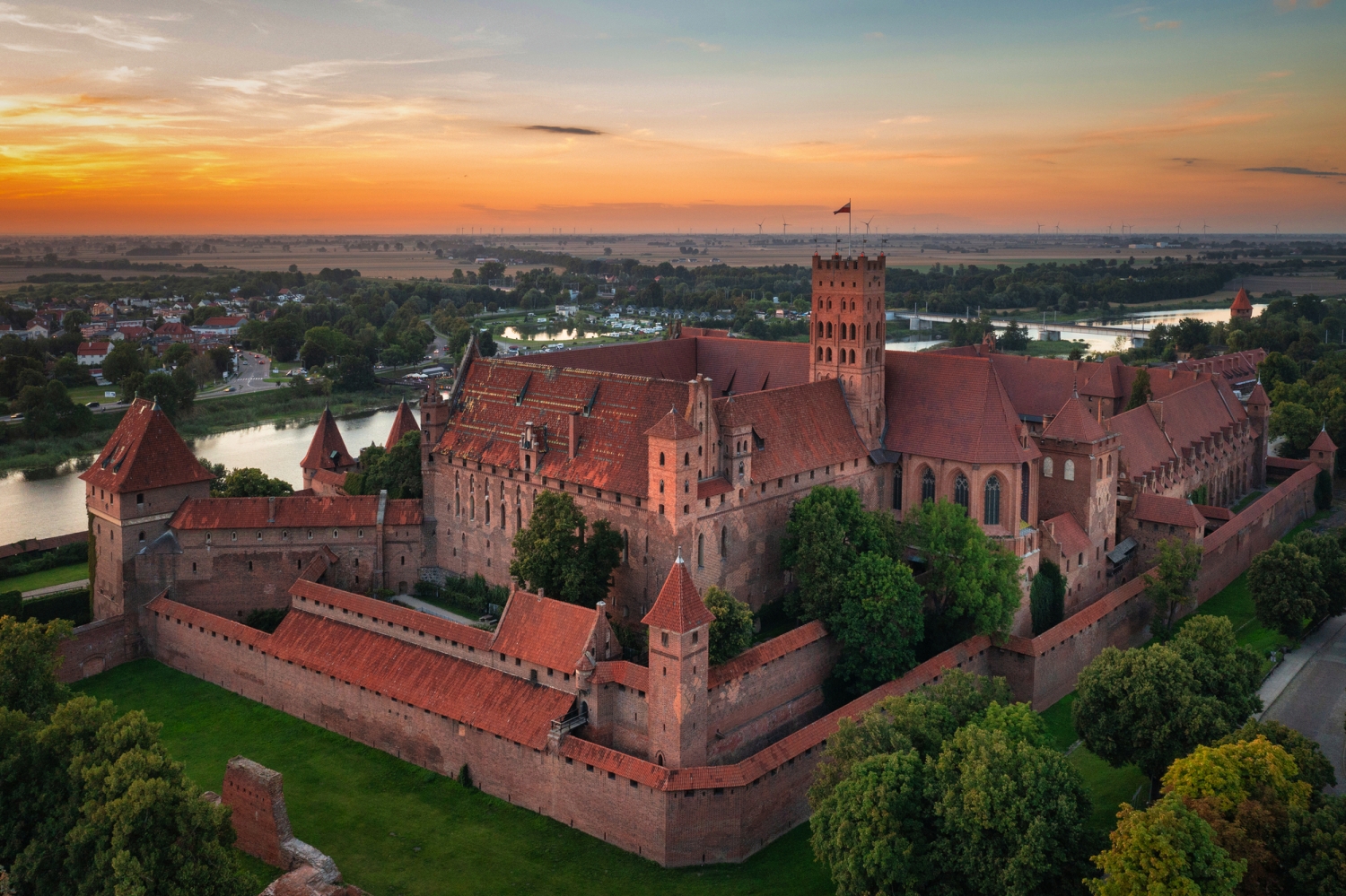 © Patryk Kosmider - stock.adobe.com Luftaufnahme einer mittelalterlichen Burg bei Sonnenuntergang, umgeben von Bäumen und Wasser.