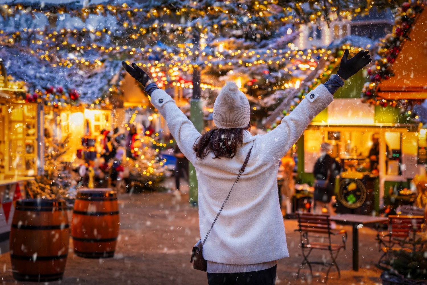 © moofushi - stock.adobe.com Person auf Weihnachtsmarkt, Hände hoch, umgeben von Lichtern und Dekorationen.