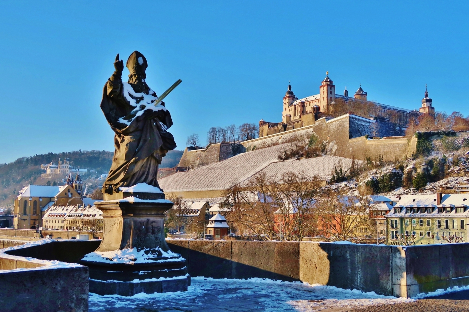 Statue auf einer Brücke vor einer verschneiten Festung und Stadt im Hintergrund.