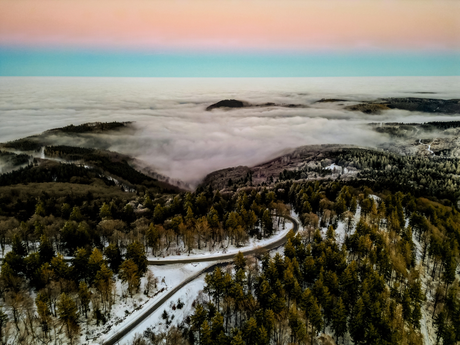 Winterlandschaft mit verschneitem Wald, kurviger Straße und Nebel in den Bergen.