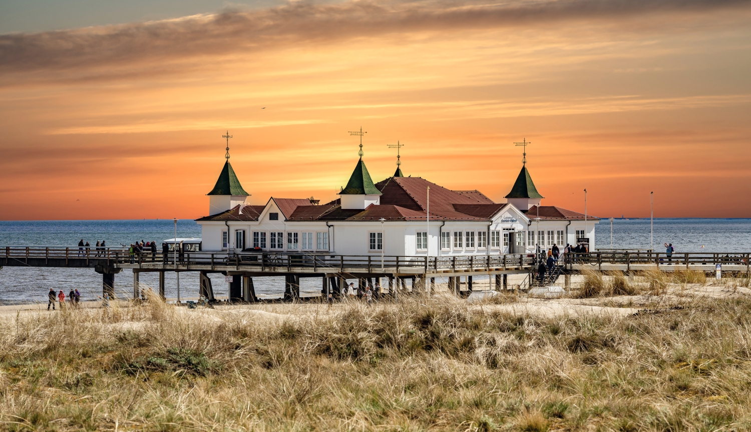 Seebrücke bei Sonnenuntergang mit grasbedeckter Düne im Vordergrund.