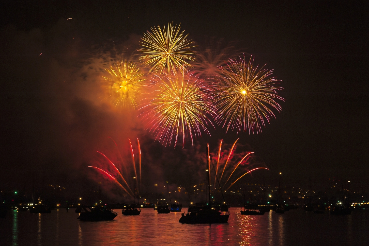 Feuerwerk über einem Wasser bei Nacht mit Booten im Vordergrund.